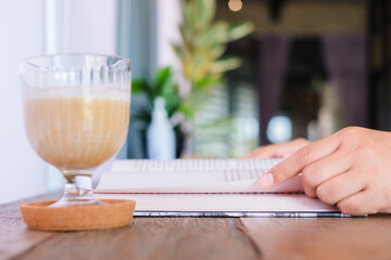 Closeup hand of a woman going to flip the page of a pocketbook on the table with a cold coffee in a...