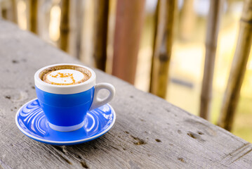 Hot cappuccino coffee in blue ceramic mug on wood bench, near the bamboo fence on background