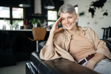 Confident positive self-sufficient woman with a short hairstyle in the interior of her house sitting on the couch, looking at the camera.