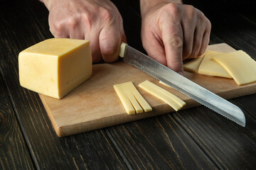 The hands of the cook with a knife cut the cheese into small pieces for tasting on a cutting board....