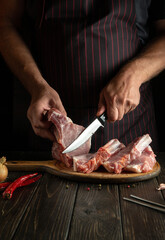 The cook cuts raw ribs with a knife on a kitchen cutting board. Barbecue grill. Idea for a restaurant or hotel menu on a dark background