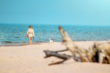 Plaża po której spaceruje Kobieta w kapeluszu, razem ze swoim  psem. Widok na morze, ocean plaże i leżący  konar drzewa.