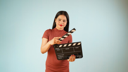 Beautiful young Asian Indian woman standing holding clapperboard, clapper board used in film making, isolated on blue color background studio portrait