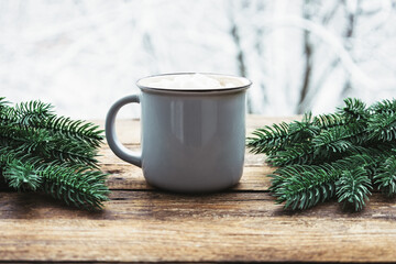 Obraz na płótnie Canvas Grey cup with chocolate or cocoa, marshmallow and fir tree decoration on wooden table against backdrop of treess with snow on cold winter day. Hot warm drink in frosty weather. Cozy composition.