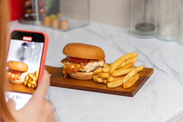 Female hands taking photo of food with mobile phone, friends using smartphones to take photos of burger and french fries on a wooden surface.
