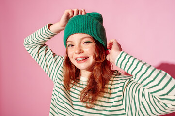 Happy smiling freckled redhead teenager girl with dental retainer, wearing trendy green beanie hat, striped long sleeve shirt, posing on pink background. 
