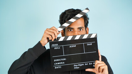 Young Asian Indian man standing holding clapperboard shocked, hiding behind clapper board used in...