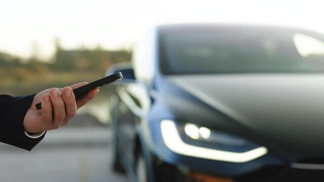 Unrecognizable elegant man using smartphone app for remote controlling of premium class Tesla Plaid car, driver distantly approaching car and opening door, smart car