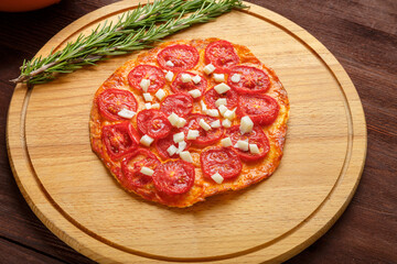 Homemade pizza with tomatoes and mozzarella on a round wooden board next to a sprig of rosemary.