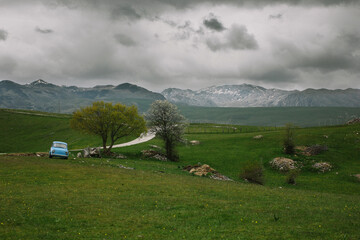 the road goes along a field with a haystack against the backdrop of mountains with trees, an abandoned old small blue car