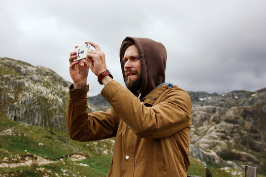a man with a beard in a brown hoodie and jacket takes pictures with an phone of a landscape in the mountains