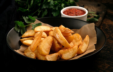 Fried potatoes in large pieces with ketchup in a black plate on a rustic wooden background, close up