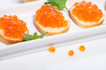 Orange caviar scattered around table and caviar on small butter rolls decorated with parsley on white background and on white rectangular plate.