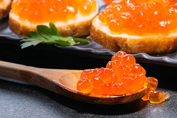 Red caviar close-up on wooden spoon and on butter buns on black plate. Caviar on spoon and on butter buns.