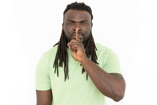 Serious African American man making silence gesture. Front view of man with dreadlocks looking at camera, asking to be quiet with his finger on lips. Silence, hush, secrecy concept