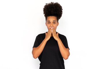 Fototapeta na wymiar Portrait of shocked young woman covering mouth with hands against white background. African American woman wearing black T-shirt looking at camera in astonishment. Surprise concept