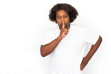 Fototapeta na wymiar Cheerful African American man shushing. Portrait of happy mature male model with dark curly hair in white T-shirt looking at camera, pressing finger on lips, asking to be quiet. Secret, gossip concept