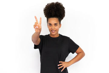 Portrait of happy young woman making victory gesture against white background. African American woman wearing black T-shirt looking at camera and smiling. Happiness and success concept