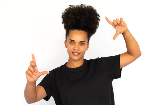 Portrait of happy young woman making frame gesture against white background. African American woman wearing black T-shirt looking at camera and smiling. Photographing concept