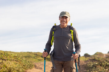 Portrait of smiling male hiker with grey hair. Man in casual clothes with hiking ammunition looking at camera, spectacular landscape in background. Hobby, nature concept