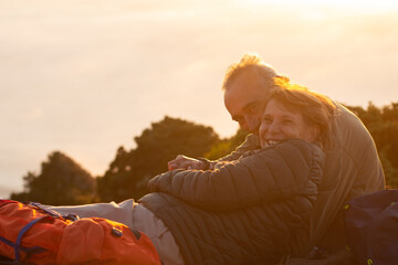 Warm elderly hikers at sunset. Man and woman in casual clothes and with ammunition sitting at peak,...