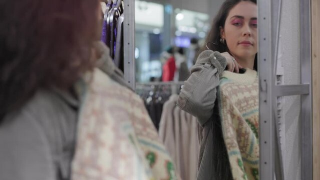 Portrait of young beautiful woman choosing sweater in store. Girl with make up trying new fashionable clothes. Concept of shopping and sale.