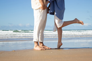 Romantic couple standing at seashore on wet sand holding hands. Slim woman in long blue shirt reaching up for kissing her boyfriend in white trousers. Fantastic sea background. Love, holiday concept