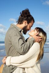 Obraz premium Romantic couple spending time at seashore on sunny day. Tall guy with man bun holding girls face in his hands, asking for kiss while happy young woman hugging her boyfriend. Romance, leisure concept