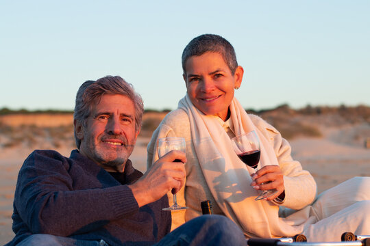 Happy senior couple drinking red wine at seashore at sunset while having picnic and enjoying warm evening at seaside. Short-haired lady and bearded man smiling at camera. Romance, leisure concept
