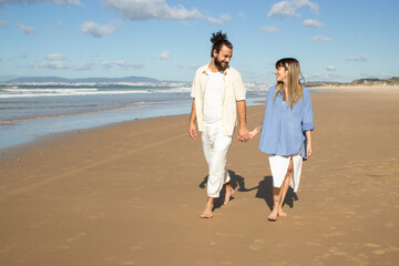 Fototapeta na wymiar Young Caucasian couple strolling along seashore holding hands and looking at each other. Tall bearded guy with man bun and blonde girl with tattoo on her arm walking barefoot. Romance, leisure concept
