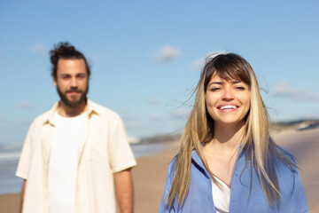 Beautiful Caucasian woman smiling while spending time at seashore with her boyfriend. Happy lady with long hair looking at camera and laughing with blurred man in background. Lifestyle, love concept