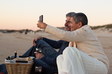 Happy senior couple having picnic at seashore in evening, drinking wine and taking selfie on mobile phone. Grey-haired man smiling while lady holding smartphone. Modern technology, romance concept