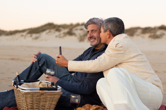 Romantic senior couple enjoying picnic at seashore in evening, drinking red wine and taking selfie. Grey-haired man smiling while lady with smartphone kissing his cheek. Leisure, romance concept - Powered by Adobe