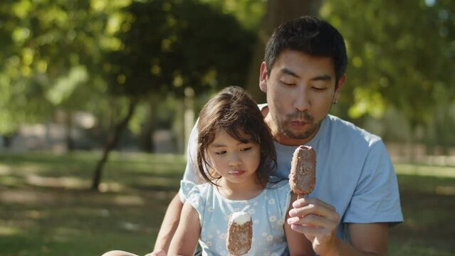 Happy Asian father and daughter eating choc ice in park. Young man sitting with little child outdoors eating ice cream. Fatherhood and childhood concept