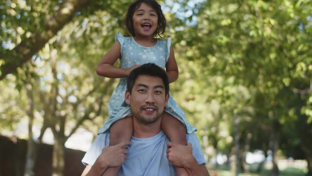 Young Asian man giving his daughter a piggyback ride in park. Happy little girl walking with father outdoors. Fatherhood and childhood concept