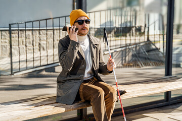 Blind person with a walking stick sits on a bench at a public bus stop uses a smartphone