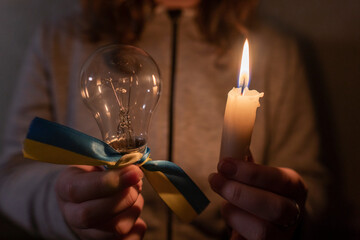 The girl holds in her hands a burning candle and an electric light bulb with a yellow-blue ribbon...