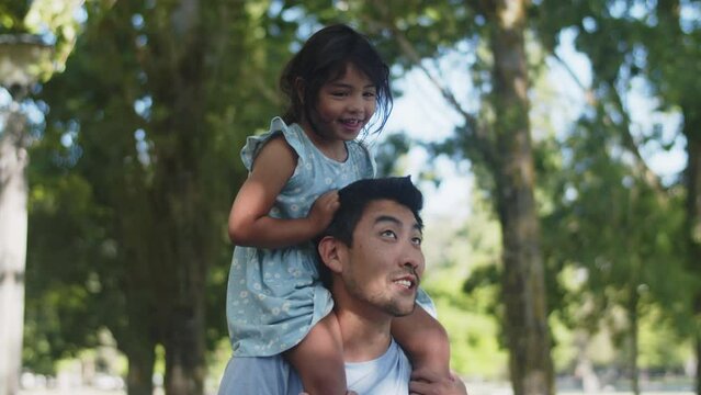 Happy Asian girl riding her father piggyback in park. Young man walking with little daughter, child laughing. Fatherhood and childhood concept