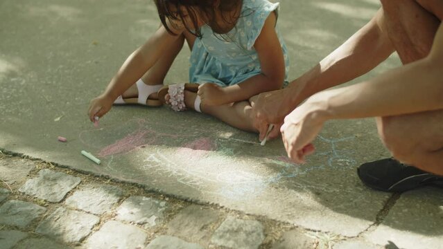 Father and child sitting on asphalt and drawing with chalks. Asian little daughter wearing blue dress walking with dad in park. Fatherhood and childhood concept