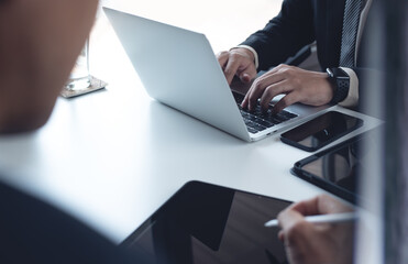 Fototapeta premium Businessman in black suit sitting at table with his colleague, working on laptop computer and digital tablet in moden office, business meeting, teamwork concept, close up