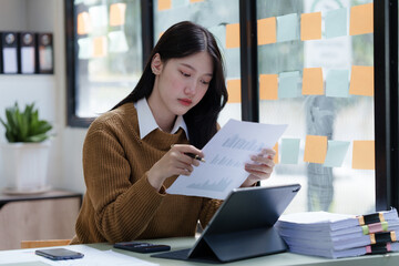 Cheerful asian businesswoman wearing sweater working at workplace female office worker during working day in office, working serious, looking at chart paper