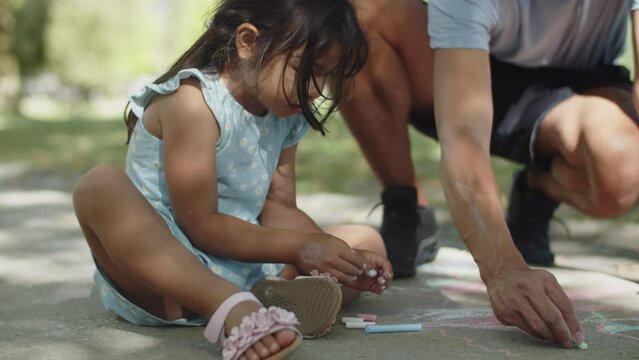 Happy Asian girl and her father drawing with colorful chalks on asphalt in park. Cute daughter wearing blue dress having leisure with her dad. Fatherhood and childhood concept