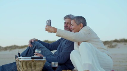 Happy senior couple having picnic at seashore in evening, drinking wine and taking selfie on mobile phone. Grey-haired man smiling while lady holding smartphone. Modern technology, romance concept. - Powered by Adobe
