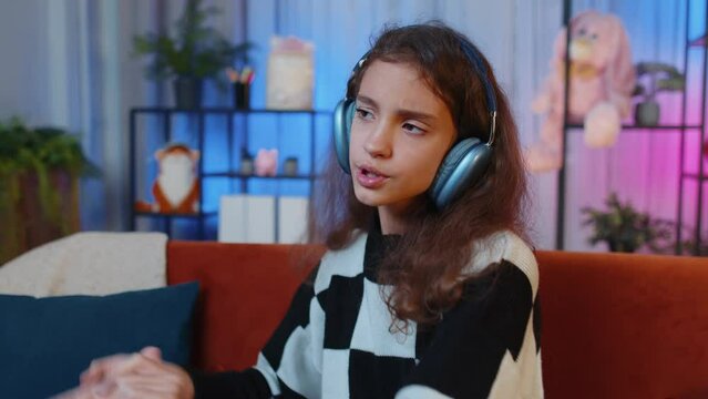 Happy overjoyed preteen girl in wireless headphones relaxing sitting on couch at night home choosing listening favorite energetic disco dancing music. People weekend leisure activities. Lifestyle