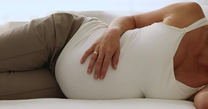 Expectant mother lying on side, touching belly, holding baby bump, feeling kicks, sleeping in bed at day time. Pregnant woman enjoying leisure, relaxation at home. Cropped shot, close up of stomach