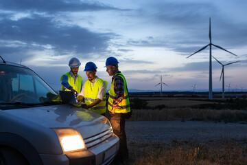 Group of engineers and maintenance workers using laptop together discussing plans in wind turbine farm at sunset.
