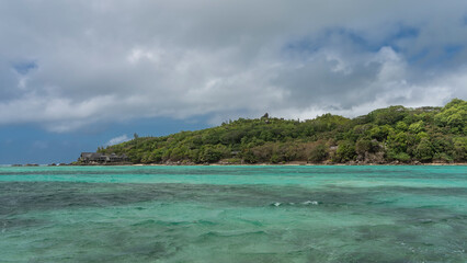 The island in the turquoise ocean is completely overgrown with tropical vegetation. The villas of the hotel are visible among the trees. Blue sky with clouds. Seychelles