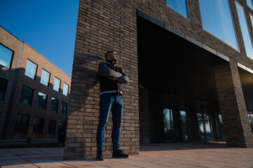 Caucasian bearded man in a bomber jacket leaned against a brick wall. 