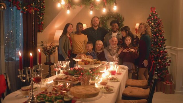 Asian woman sets timer on camera or phone. Happy multi cultural family takes group photo. They celebrating Christmas or New Year. Table with dishes and candles. Family Christmas dinner. Camera view.