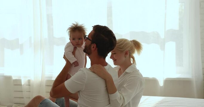 Two happy new parents cuddling baby in bed in home bedroom, playing with sweet few months infant, having fun together. Young mom and dad holding kid in arms enjoying parenthood, family leisure